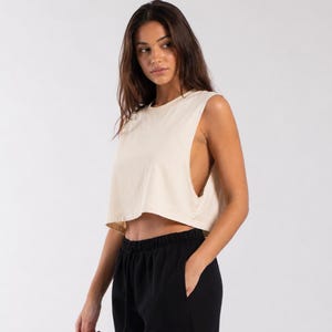 May include: A cream-colored sleeveless crop top with a crew neckline and wide arm openings, paired with black sweatpants featuring an elastic waistband and a drawstring. The top is constructed from a solid fabric and sits above the waistline.