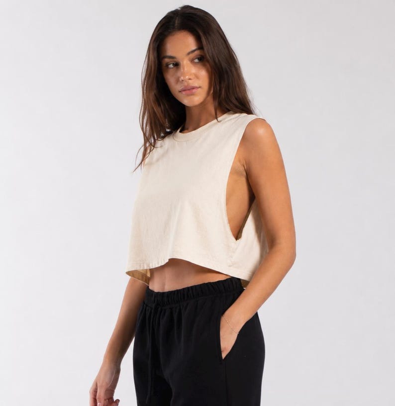 May include: A cream-colored sleeveless crop top with a crew neckline and wide arm openings, paired with black sweatpants featuring an elastic waistband and a drawstring. The top is constructed from a solid fabric and sits above the waistline.