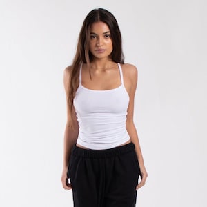 May include: A white tank top with thin straps. The top is form-fitting and has a scoop neckline. The model is wearing black sweatpants. The background is plain white.