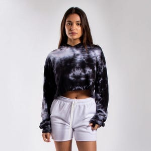 Pode incluir: Um moletom curto de manga comprida com padrão tie-dye preto e branco, com capuz e punhos canelados, combinado com shorts brancos lisos que possuem cós elástico e um cordão branco pendurado no centro da parte frontal.