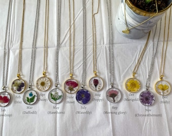 May include: Twelve gold and silver necklaces with round glass pendants featuring pressed flowers. Each pendant is labeled with a month and the corresponding flower name, such as January (Snowdrop), February (Iris), March (Daffodil), and so on.