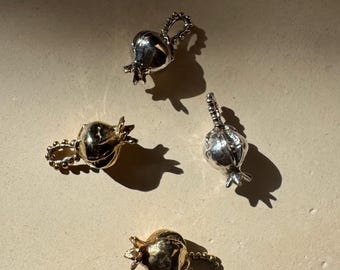 May include: Four small, metallic pomegranate charms. Two are silver-toned, and two are gold-toned. Each charm has a loop for attaching to a chain or bracelet. The charms are scattered on a light-colored surface, with shadows cast by sunlight.