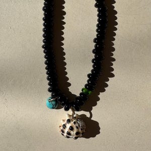May include: A black beaded necklace with a small turquoise bead and a seashell pendant. The seashell is white with brown spots. The necklace is approximately 18 inches long.