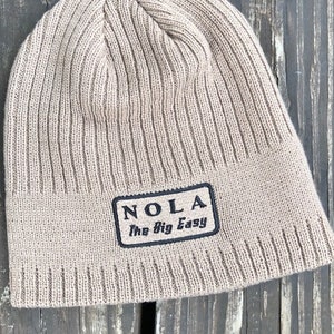 May include: A beige knitted beanie with a black patch that reads "NOLA The Big Easy".