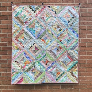 May include: A handmade quilt with a patchwork design, featuring a variety of colorful fabric scraps. The quilt is composed of numerous squares and diagonal lines, with a pink border. The quilt is hanging against a brick wall.