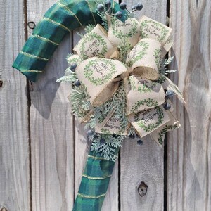 May include: A green and gold plaid candy cane Christmas wreath with a burlap bow and sprigs of greenery. The bow has multiple tags that say "Merry Christmas".