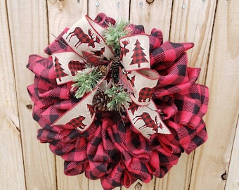 May include: A red and black buffalo plaid wreath with a brown and white ribbon featuring moose and bear silhouettes and a small pine tree. The ribbon is tied into a bow with a pine sprig and pine cones.