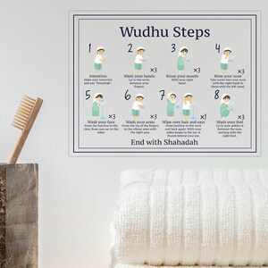 Op de afbeelding: Een rechthoekig wandbord getiteld Wudhu Steps met acht genummerde illustraties die het rituele wasproces tonen, inclusief tekstinstructies voor elke stap, boven twee gevouwen witte handdoeken en een bruine marmeren tandenborstelhouder.