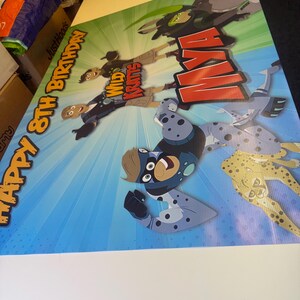 May include: A colorful birthday banner featuring the Wild Kratts characters. The banner has a blue and green background with a red and white "Happy 8th Birthday" message. The characters are depicted in their animal suits, including a cheetah, a leopard, and a monkey.