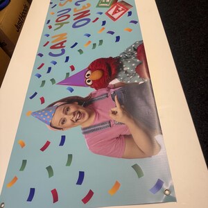 May include: A blue banner with a red and white Sesame Street character, Elmo, and a woman wearing a pink shirt and a blue overall. The banner has the text "Can you say one?" and colorful confetti.