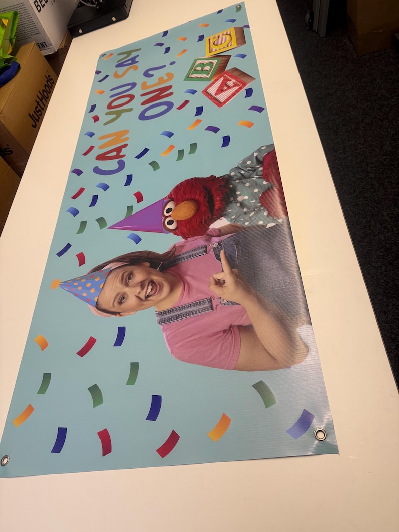 May include: A blue banner with a red and white Sesame Street character, Elmo, and a woman wearing a pink shirt and a blue overall. The banner has the text "Can you say one?" and colorful confetti.