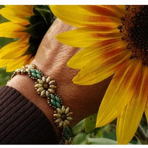 May include: A beaded bracelet with a floral design in shades of gold, green, and white. The bracelet is worn on a wrist against a backdrop of a bright yellow sunflower.
