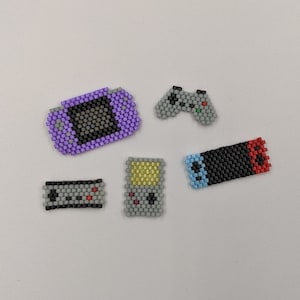 Puede incluir: Cinco mini mandos y consolas de videojuegos de cuentas. Las consolas son moradas, grises y negras con detalles rojos y azules. Los mandos son grises con detalles negros y rojos.