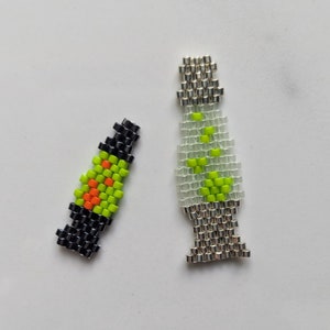 以下が含まれることがあります： 2つのビーズの溶岩ランプのイヤリング。1つは黒、オレンジ、緑です。もう1つはシルバーとクリアで、緑と黄色のビーズが付いています。イヤリングは白い表面にあります。