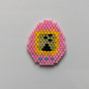 May include: A pink beaded egg with a yellow face and black eyes. The egg has three blue beads at the bottom.