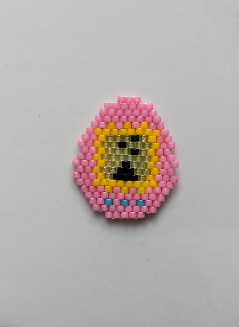 May include: A pink beaded egg with a yellow face and black eyes. The egg has three blue beads at the bottom.