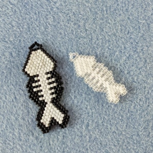 Puede incluir: Dos abalorios de pescado de cuentas, uno blanco y negro y otro blanco. El pez blanco y negro tiene un diseño de esqueleto.