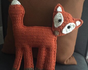 May include: A crocheted fox plush toy with orange fur, white facial features, and brown feet. The fox is sitting on a brown pillow.