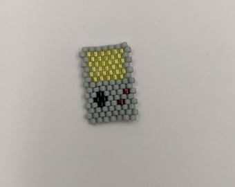 Peut inclure: Un charm fait à la main en perles en forme de console de jeu rétro. Le charm présente un corps gris avec un écran jaune, des détails noirs pour l'écran et des accents rouges. Le charm est fait de petites perles colorées.