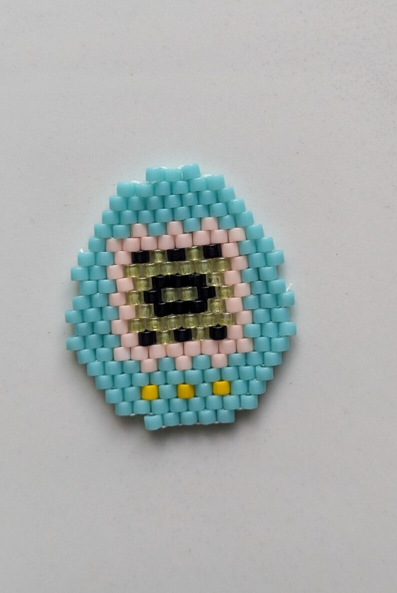 May include: A blue and yellow beaded egg with a pink and black face. The face has a black eye and a yellow mouth.