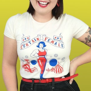 Könnte beinhalten: Ein weißes T-Shirt mit einem rot-weiß-blauen Motiv einer Frau, die Gewichte hebt. Das Motiv lautet "Certain Female" mit Sternen auf beiden Seiten. Die Frau trägt ein rot-weiß gestreiftes Tanktop und blaue Shorts. Das T-Shirt hat einen roten Gürtel.