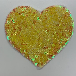 May include: A yellow heart-shaped patch made of iridescent sequins. The sequins are arranged in a circular pattern, creating a shimmering effect.