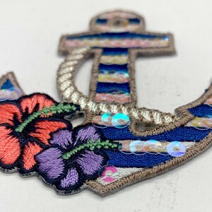 Puede incluir: Parche bordado de ancla con rayas azules y blancas, decorado con lentejuelas iridiscentes y dos flores de hibisco bordadas, una rosa y una morada.