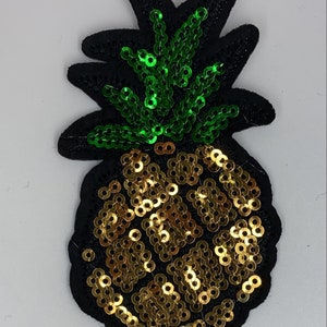 Può includere: Una toppa a forma di ananas dorata e verde con paillettes. L'ananas è bordato di feltro nero.