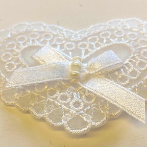May include: White lace heart with a white satin bow and gold beads in the center.