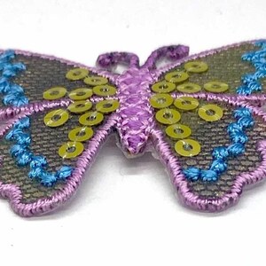 May include: A purple and green butterfly applique with yellow sequins. The butterfly has a detailed embroidered design and a pink center.