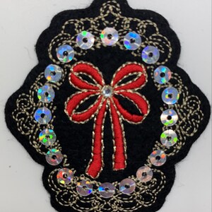 May include: A black felt applique with a red bow and gold embroidery. The applique is surrounded by a circle of iridescent sequins.