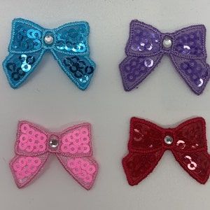 May include: Four fabric bows with rhinestones in the centre. The bows are in blue, purple, pink, and red.