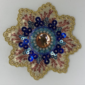 May include: A gold star-shaped fabric applique with a central circle of blue and turquoise sequins, surrounded by a ring of smaller blue sequins and a large, clear, faceted, orange rhinestone.
