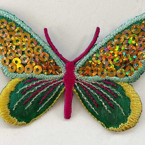 Op de afbeelding: Een groene vlinderapplicatie met gouden en iriserende pailletten, roze stiksels en een gele omtrek.