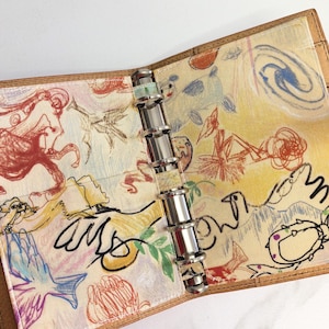 May include: Open leather-bound planner with a silver binder mechanism. The interior lining features a colourful, whimsical design with drawings in red, blue, yellow, and green. The planner is open, showcasing the artistic inner pages.