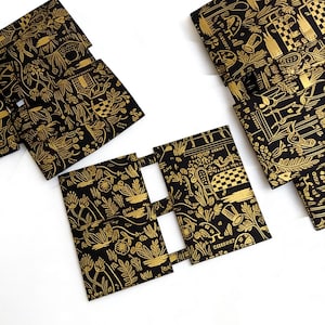 Könnte beinhalten: Mehrere schwarze Stoffartikel mit einem goldenen Muster, das botanische und architektonische Elemente zeigt. Die Artikel scheinen gefaltet oder geformt zu sein, möglicherweise zur Aufbewahrung oder Organisation. Das goldene Design hebt sich stark vom schwarzen Hintergrund ab.