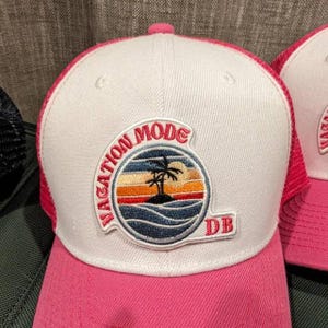 Può includere: Cappellino da camionista bianco e rosa con una toppa che dice "Vacation Mode" e una grafica di una palma. Il cappello ha anche un piccolo logo "DB" sul lato.