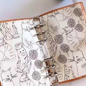 May include: Open planner with a white fabric interior featuring a repeating pattern of sketched cats and yarn balls in black. The planner has a brown leather-like cover and silver metal rings.