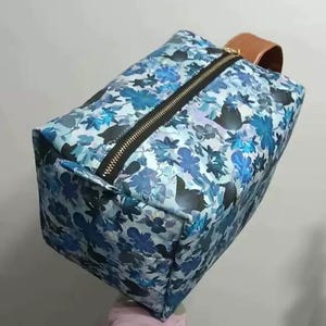 May include: A rectangular cosmetic bag with a blue floral pattern. The bag has a black zipper and a brown leather handle. The floral design features various shades of blue and white. The bag is designed to hold makeup and other beauty products.