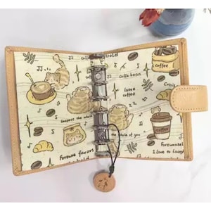 May include: Open beige planner with a coffee and cat theme. The interior features a cream-colored fabric with illustrations of coffee cups, beans, cats, and text. The planner has a silver binder and a beige leather closure.
