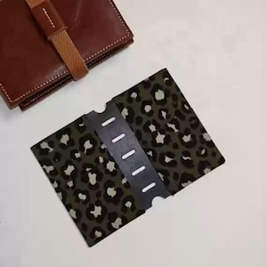 Puede incluir: Una cartera de cuero marrón con una correa beige y un tarjetero con estampado de leopardo verde y blanco. El tarjetero tiene una correa negra con recortes blancos. Los artículos están sobre una superficie blanca.