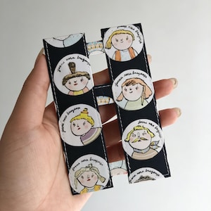 May include: A black fabric bookmark featuring a repeating pattern of white circular illustrations. Each circle contains a hand-drawn character portrait and the handwritten text Joana Rosa Bragança. The bookmark has white stitched edges.