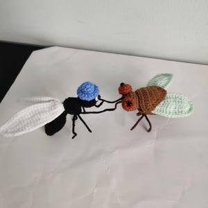 May include: Two crocheted insects are shown on a white surface. One is a black fly with white wings and a blue hat. The other is a brown insect with green wings. The insects appear to be interacting, possibly in a playful manner.