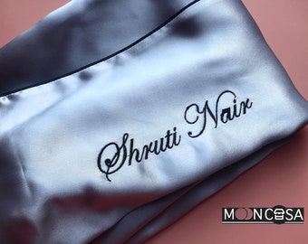 May include: A light blue satin sleep mask with black trim and the name "Shruti Nair" embroidered in black cursive script. The mask is folded in half and the brand name "MOONCSA" is visible in the bottom right corner.