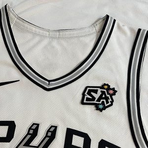 May include: White basketball jersey with black and silver trim. The jersey has a black and white logo on the front that says "Spurs" and a black patch with the letters "SA" and colourful stars on the right side.