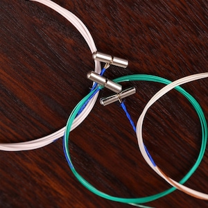 Könnte beinhalten: Ein Satz von vier Gitarrensaiten, zwei sind weiß, eine ist blau und grün und eine ist braun. Die Saiten sind um Metall-Stimmwirbel gewickelt.