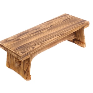 May include: A wooden bench with a natural brown finish. The bench has a simple design with a rectangular top and two legs on each side. The legs are angled slightly inward.