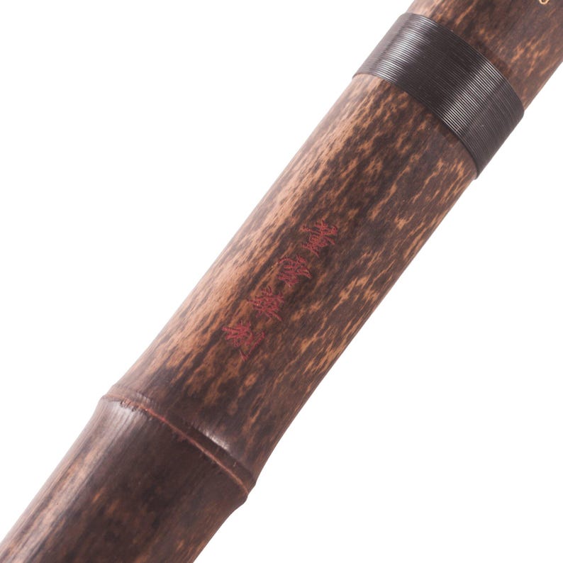 May include: Close-up of a bamboo flute with a dark brown finish and a black band. The bamboo has a natural grain pattern and red text is visible on the surface. The flute is angled diagonally against a white background.