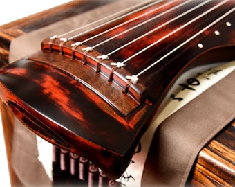 May include: A close-up of a traditional Chinese zither, a guzheng, with a red-brown finish and white strings. The instrument is resting on a brown fabric surface.