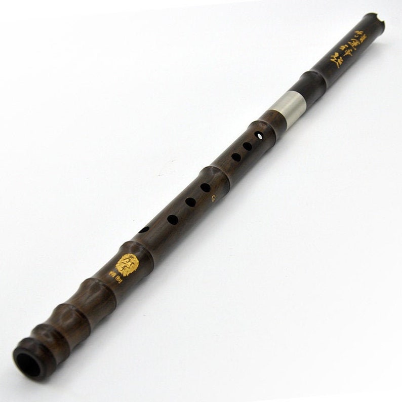 Puede incluir: Una flauta china de madera marr&oacute;n oscuro con una junta de metal plateado y letras doradas. La flauta tiene una serie de agujeros a lo largo del instrumento.